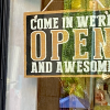 Yellow and black sign in business window saying Come in, we're open and awesome - Photo by Amber Ankerholz